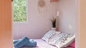 Een slaapkamer met een bed, kussens, een dromenvanger, een plant in pot en een raam.