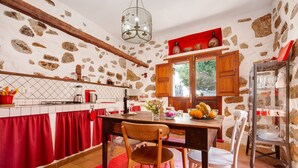 Una cocina rústica con paredes de piedra, vigas de madera, salpicadero de baldosas rojas y una zona de comedor con mesa y sillas de madera.