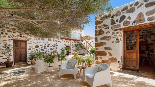 Un patio de paredes de piedra con dos sillones blancos, plantas en macetas y una puerta de madera.