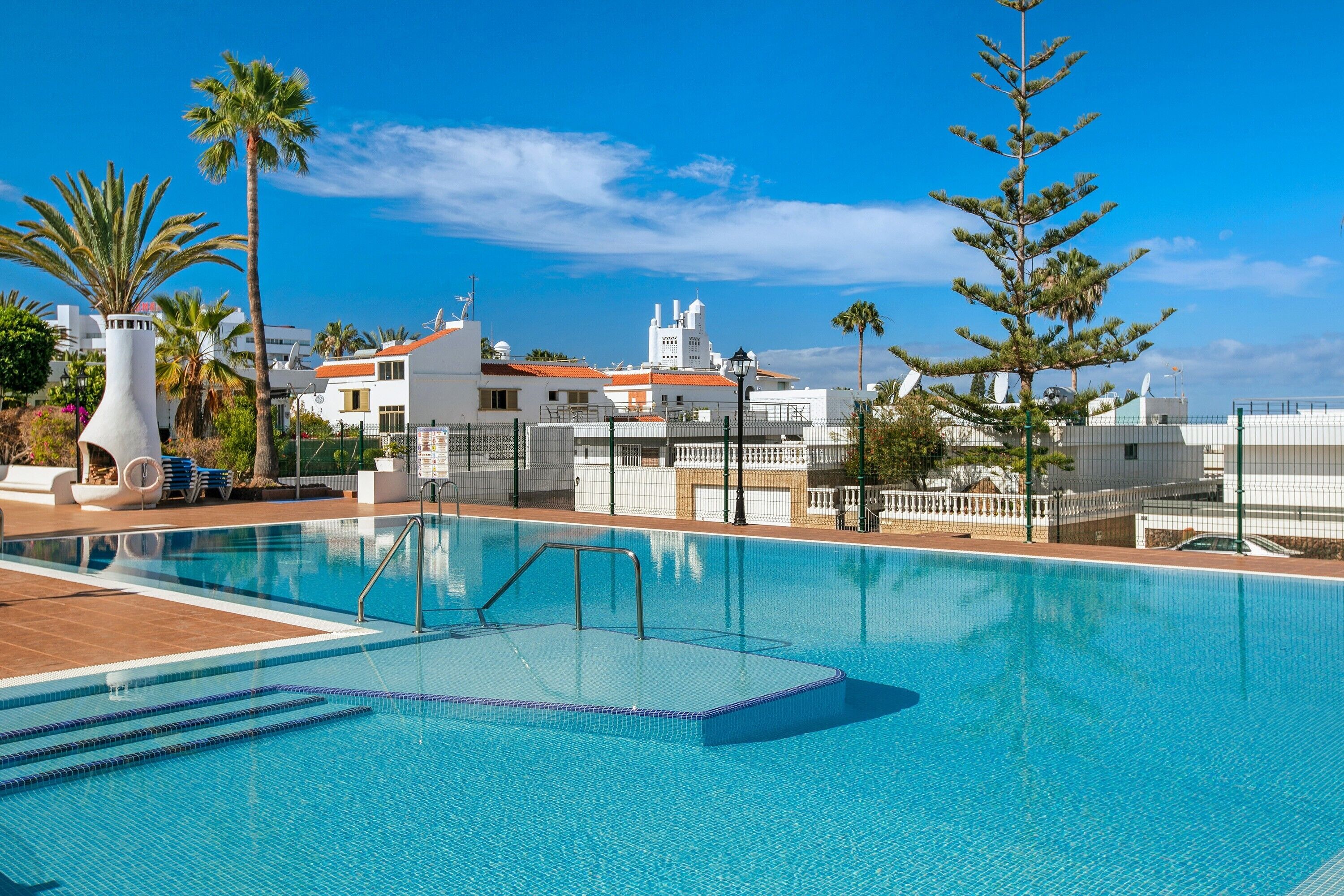 A swimming pool with clear blue water, surrounded by palm trees and a white building with a red roof.