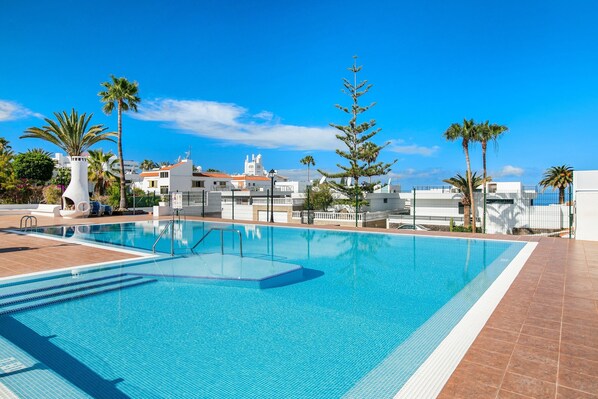 A clear blue swimming pool with a tiled deck, surrounded by palm trees and white buildings.