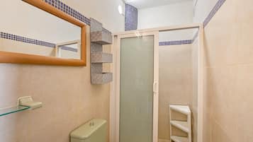 A bathroom with a glass shower enclosure, a toilet, a sink with a glass shelf, and a mirror with a tiled border.