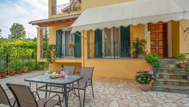 A patio with a table and chairs, potted plants, and a building with a balcony. - Apartment 'Kallithea House in Gouvia' with Mountain View, Garden & Wi-Fi (Corfu)