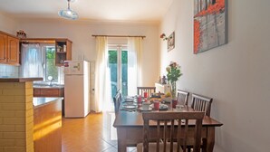 A kitchen with wooden cabinets, a refrigerator, a dining table with chairs, and a window with curtains. - Apartment 'Kallithea House in Gouvia' with Mountain View, Garden & Wi-Fi (Corfu)