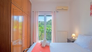 A hotel room with a bed, a nightstand, a lamp, a window with curtains, and a view of greenery outside. - Apartment 'Kallithea House in Gouvia' with Mountain View, Garden & Wi-Fi (Corfu)