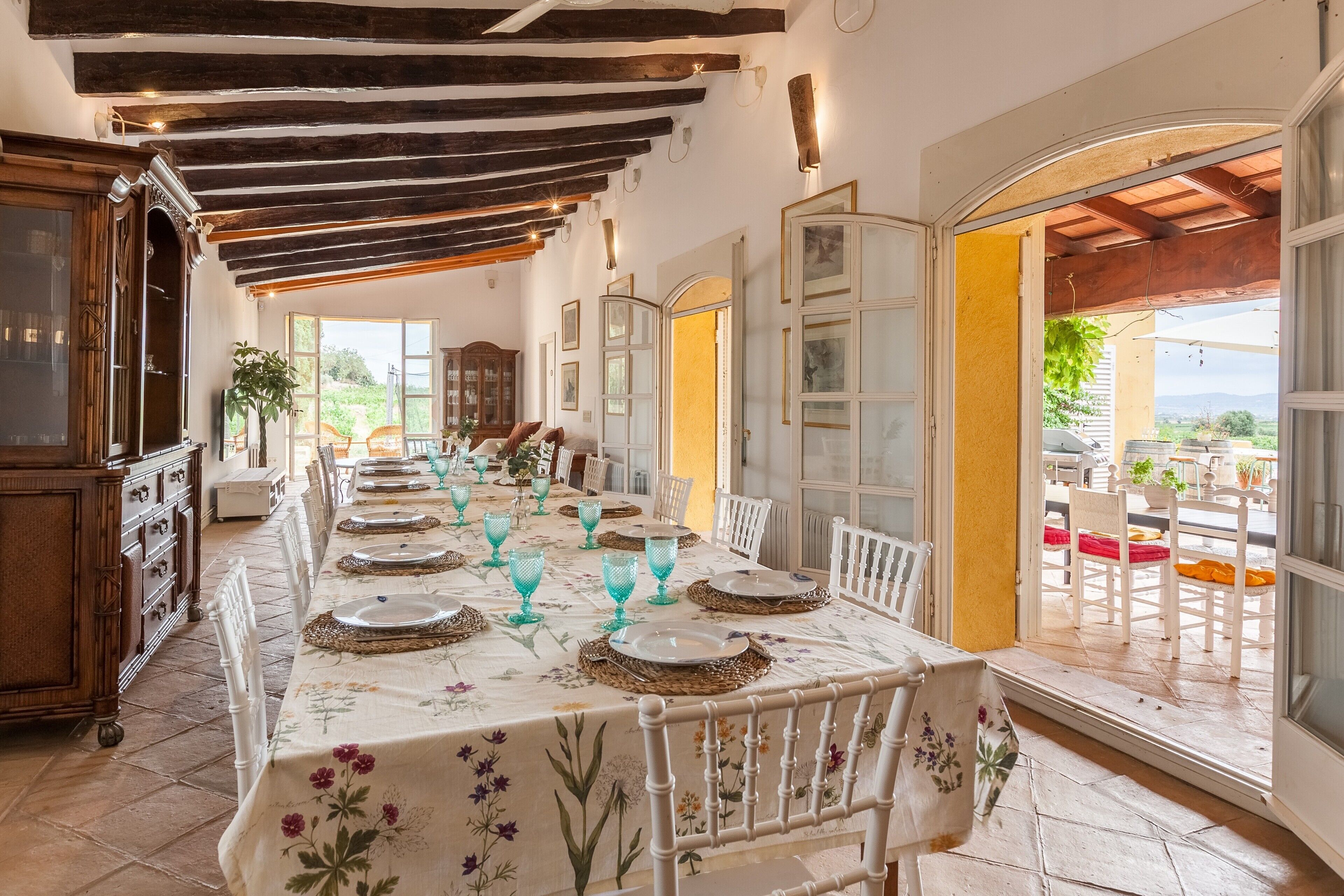 Un comedor con una larga mesa dispuesta para una comida, vigas de madera en el techo y vistas a través de un ventanal abierto.