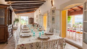 Un comedor con una larga mesa dispuesta para una comida, vigas de madera en el techo y vistas a través de un ventanal abierto.