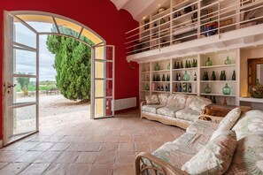 An open door leading to a patio with a view of greenery, a wicker sofa, and a wine rack with bottles. - Lidia Rural House for Groups: Pool, Views & Unique Experiences (Sant Martí Sarroca)