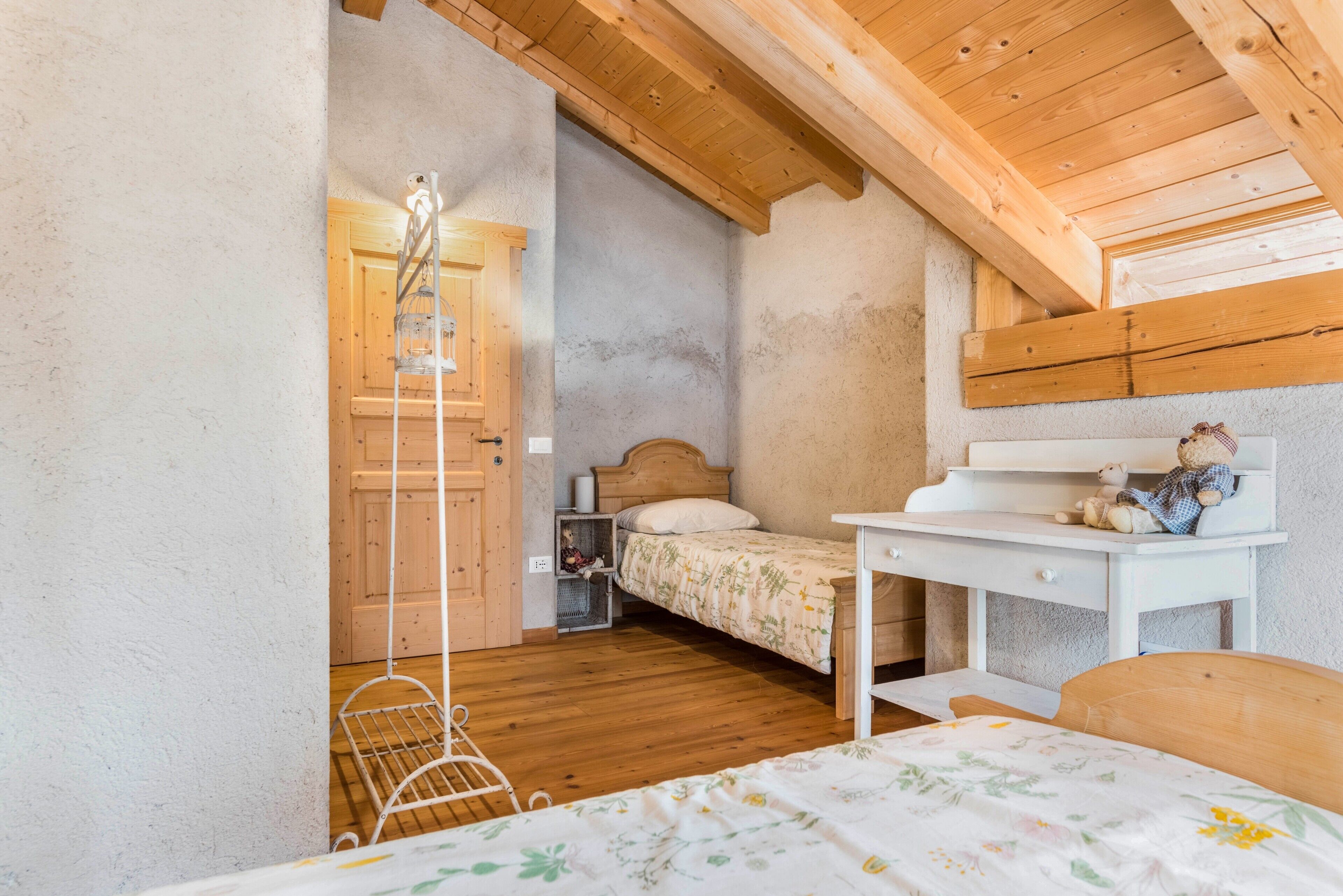 A bedroom with a wooden ceiling, a bed with a floral bedspread, a desk with a teddy bear, and a white metal chair.