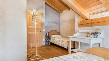 A bedroom with a wooden ceiling, a bed with a floral bedspread, a desk with a teddy bear, and a white metal chair.