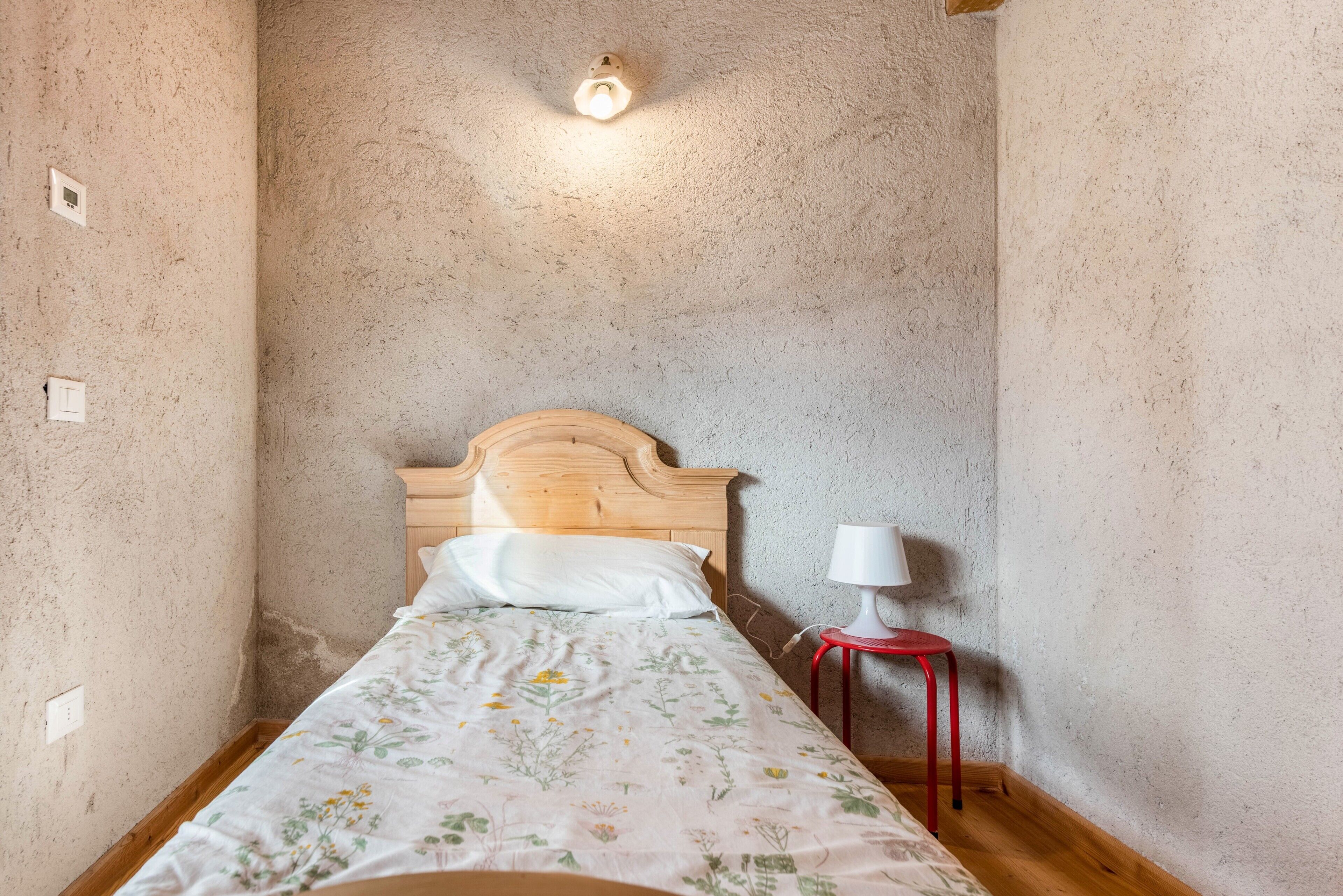Ein kleiner, schmaler Raum mit einem hölzernen Bett, einem Nachttisch mit einer weißen Lampe und einer strukturierten Wand.