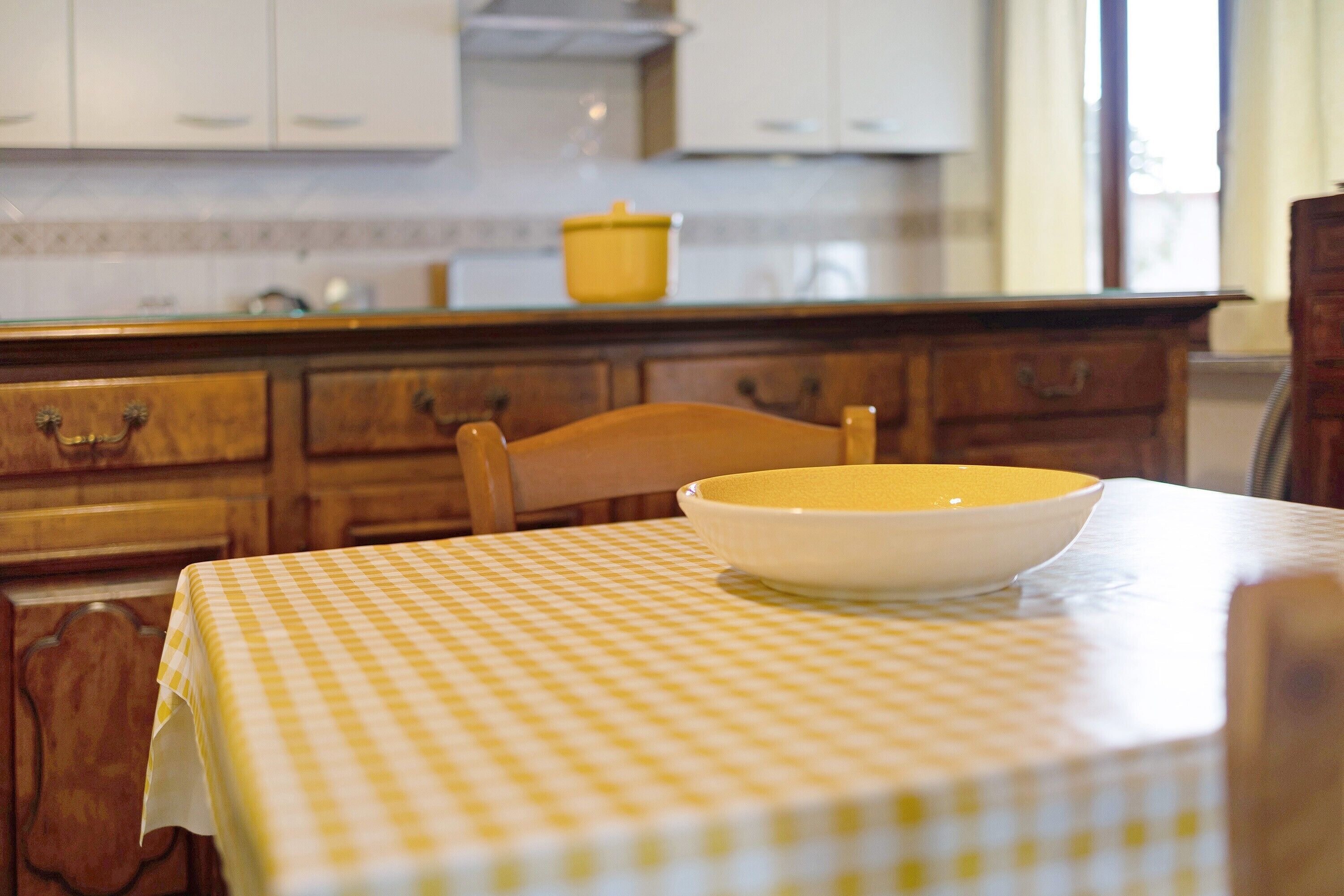 Une cuisine avec des meubles de rangement en bois, un bol jaune posé sur une table et une nappe à carreaux.