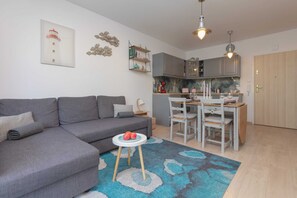 Una sala moderna con un sofá gris, una mesita redonda con manzanas, un tapete azul estampado, una cocina con gabinetes grises e iluminación colgante.