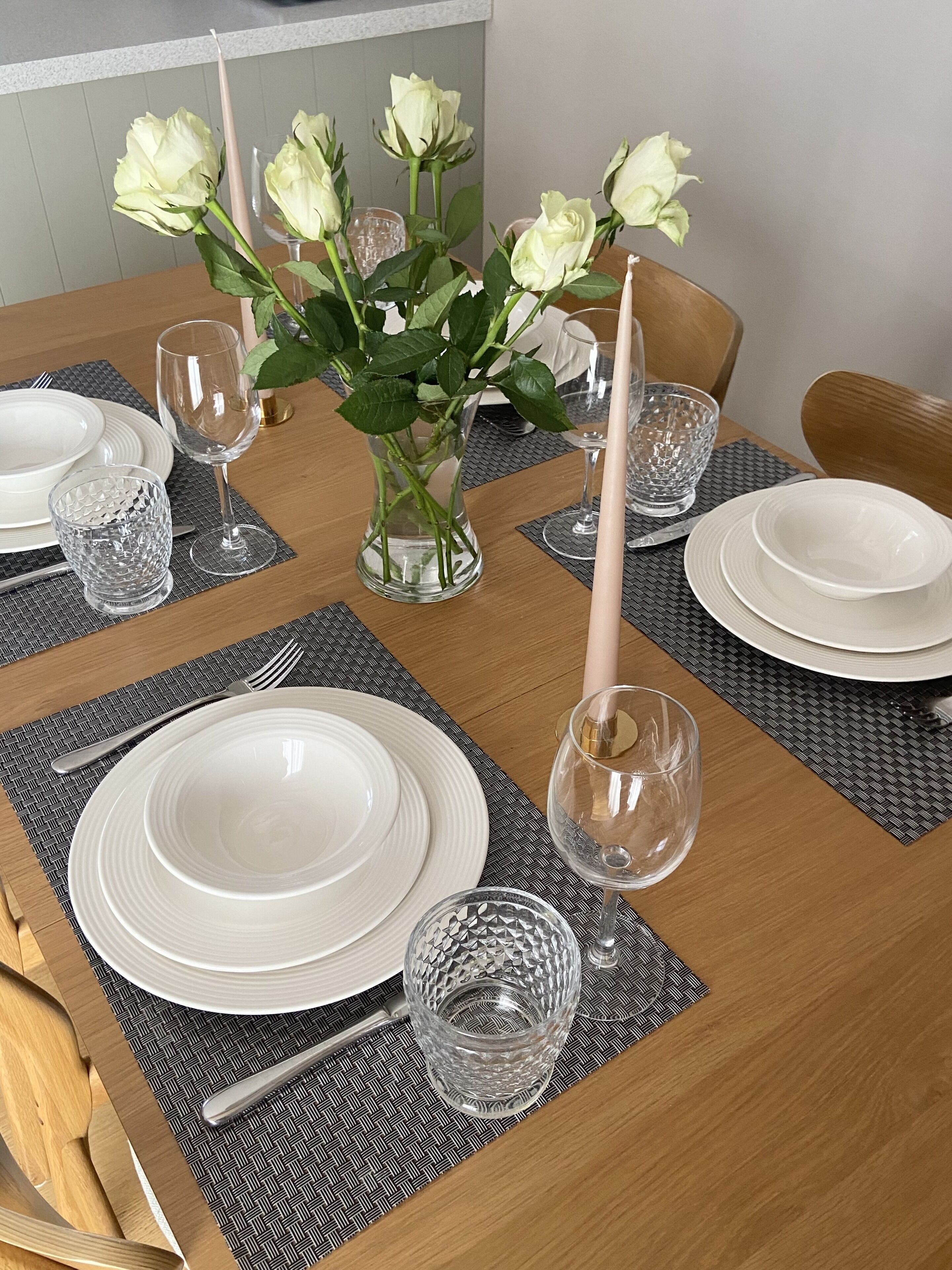 Una mesa de comedor dispuesta con platos blancos, cristalería transparente y un florero con rosas blancas.