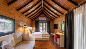 A cozy cabin interior with a wooden ceiling, a bed with a mountain-themed blanket, a sofa with patterned cushions, a desk with a kettle, and a TV. - Dolce Vita Luxury Bungalows (Bodrum)