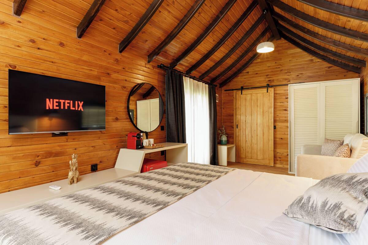 Una habitación de madera con una cama, un televisor mostrando 'NETFLIX', un escritorio, un espejo y una puerta corredera.