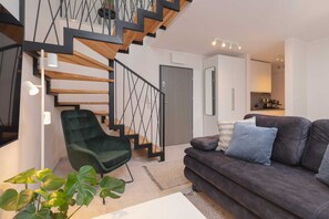 A modern living room with a staircase, a green armchair, a dark sofa, and a kitchen in the background.