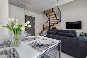 A modern living room with a dining area, a grey sofa, a TV mounted on the wall, and a spiral staircase.