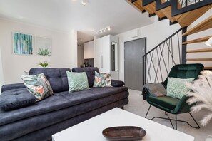 A modern living room with a dark grey sofa, a green armchair, a white coffee table, and a staircase with wooden railings.