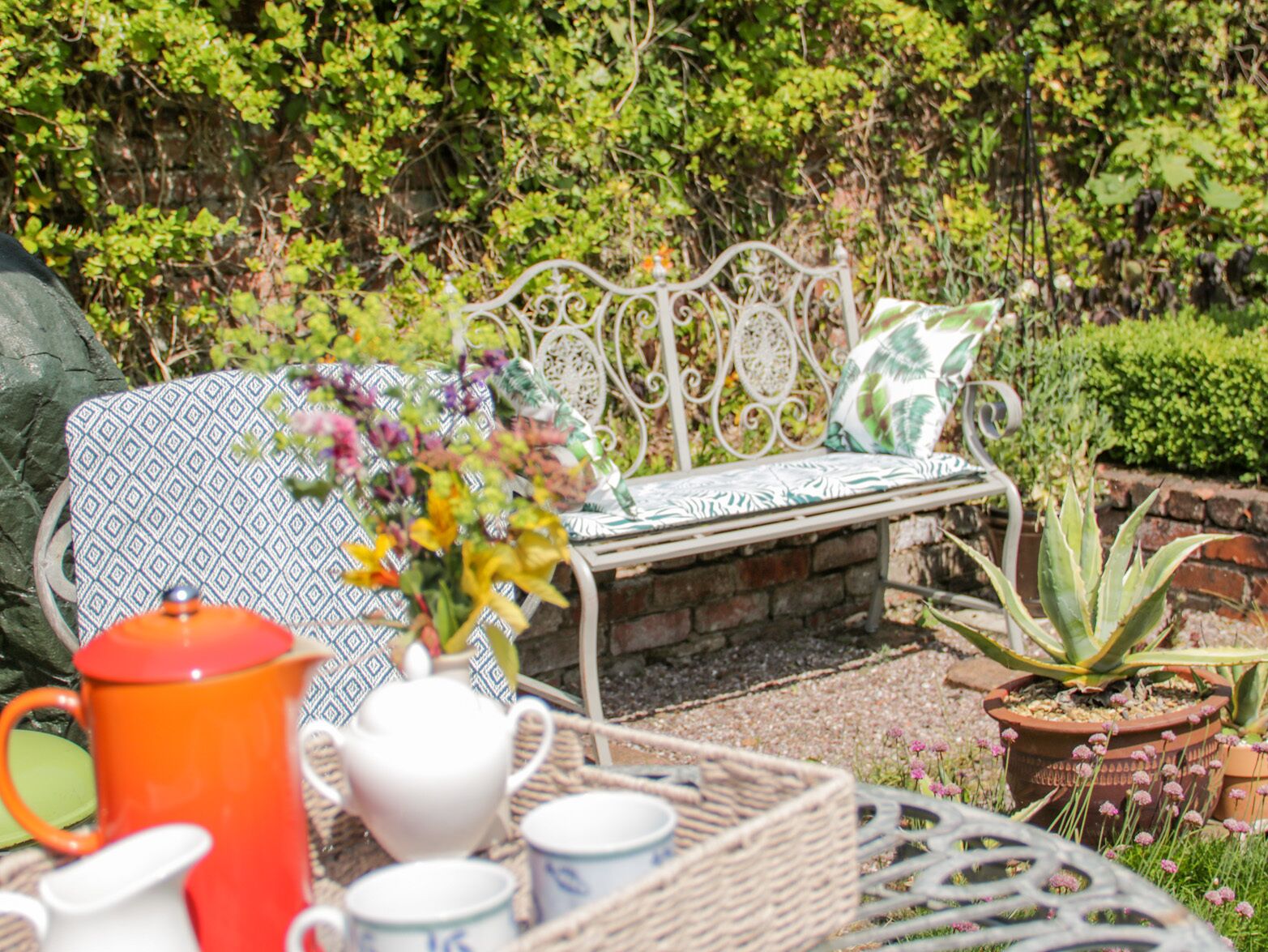 A garden setting with a metal bench, a wicker table with tea set, and potted plants.