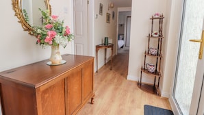 A hallway with a wooden cabinet, a mirror, a vase with flowers, a small table, and a shelf with decorative items. - Bryn Offa Cottage (Holywell)