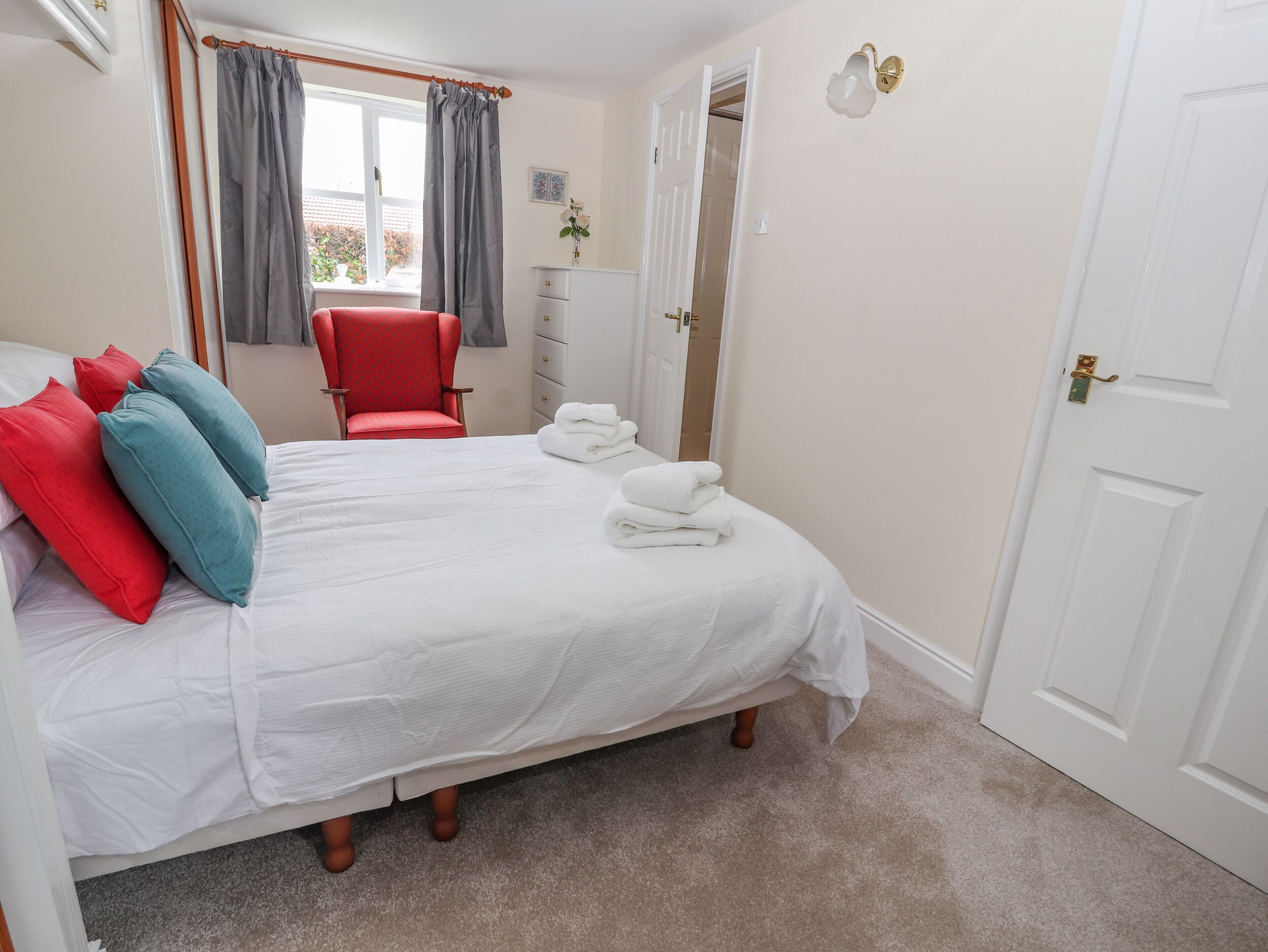 A neatly made bed with white linens and a few pillows, a red chair, a dresser, and a door with a handle.