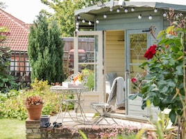A small, open-sided structure with a table and chairs, surrounded by potted plants and flowers.