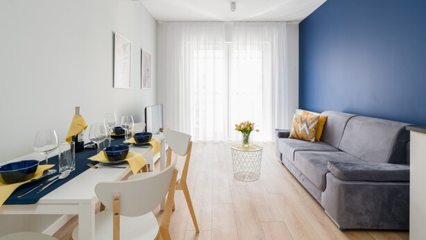 A modern living room with a dining area, featuring a sofa, a dining table set with bowls and glasses, and a vase with flowers on a side table.