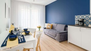A modern living room with a grey sofa, a dining table set with bowls and cutlery, a coffee machine on the counter, and a window with sheer curtains.