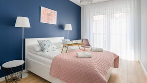 A bedroom with a bed, bedside lamps, a bedside table, a chair, a window with sheer curtains, and a wall painting.