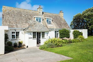 A white building with a stone roof, a front door, and a small garden area with potted plants. - Thalassa (Wadebridge)