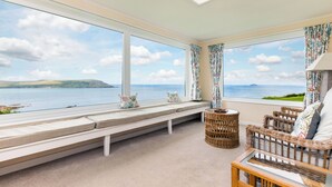 Habitación con amplios ventanales que ofrecen vistas al mar y a las colinas. Hay un banco junto a los ventanales, mobiliario de mimbre y una pequeña mesa.