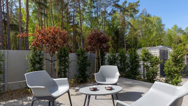 An outdoor patio with two white chairs and a small table, surrounded by trees and a wooden deck.
