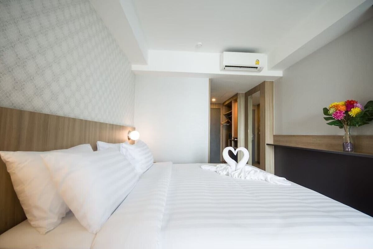 Quarto de hotel com uma cama bem arrumada, ar-condicionado fixado na parede e um vaso com flores em uma mesa de cabeceira.