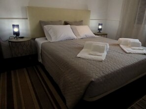 Un lit bien fait, recouvert d’une courtepointe matelassée, avec deux oreillers et deux serviettes pliées posées sur le lit. De chaque côté du lit, on trouve une table de chevet sur laquelle est posée une lampe.