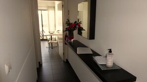 Un couloir étroit avec un comptoir noir, un miroir et un flacon de lotion. On aperçoit une chaise et une plante au fond.