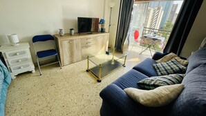 A living room with a sofa, a desk with a TV, a chair, and a window with a view of buildings.