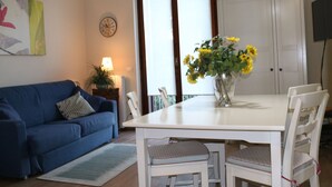 A living room with a blue sofa, a white dining table with chairs, a vase with sunflowers, a clock on the wall, and a cabinet. - Holiday apartment Two room apartment with pool (Lazise)