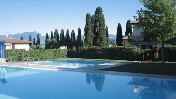 A clear swimming pool with metal ladders, surrounded by neatly trimmed hedges and tall trees. - Holiday apartment Two room apartment with pool (Lazise)