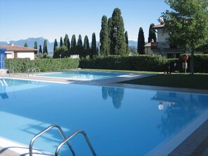 A clear swimming pool with metal ladders, surrounded by neatly trimmed hedges and tall trees. - Holiday apartment Two room apartment with pool (Lazise)