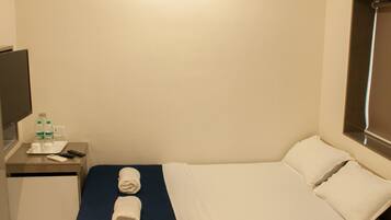 A hotel room with a bed, a desk with a flat-screen TV, a small table with a bottle of water, and a ceiling light.