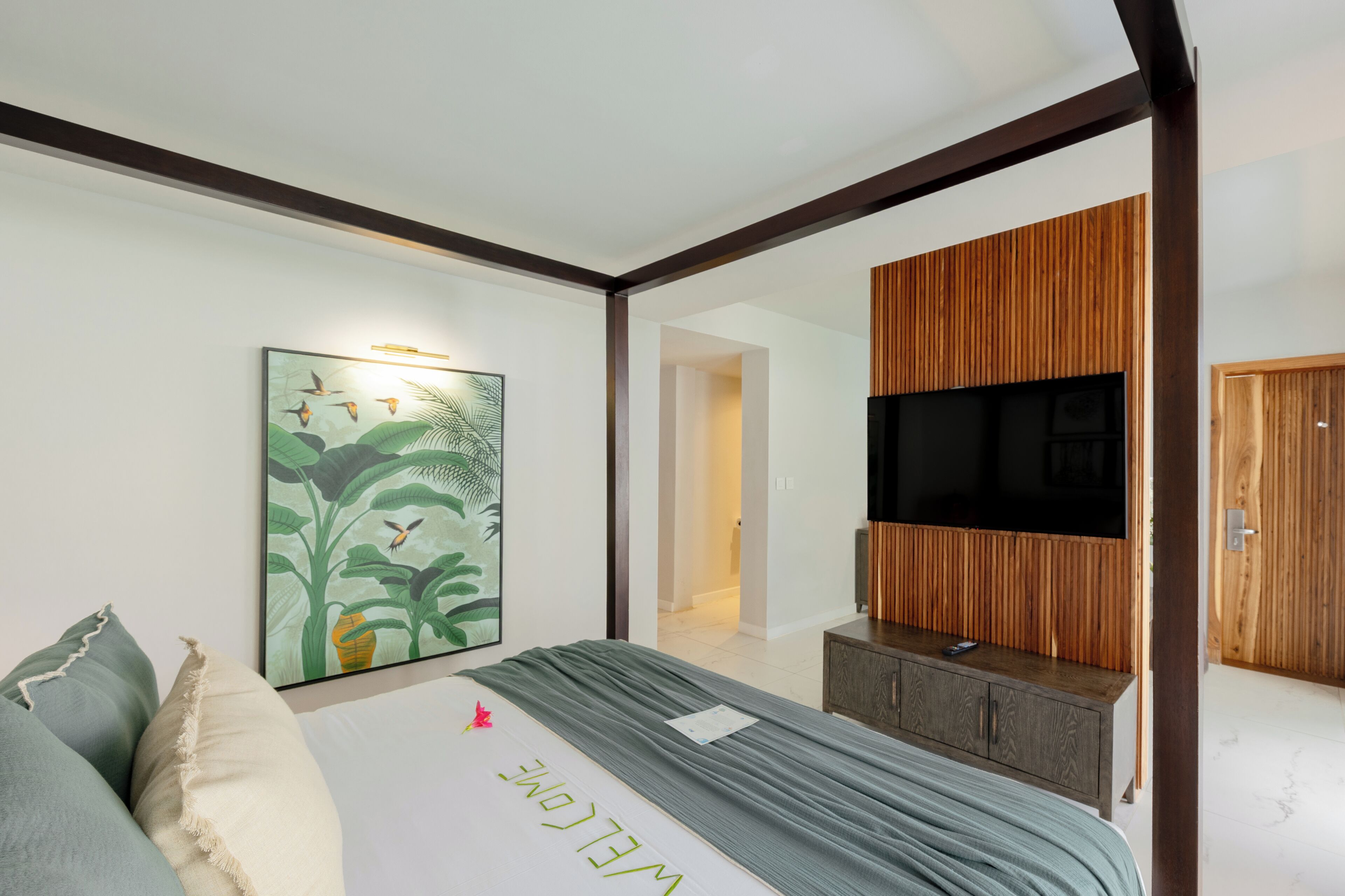 A bedroom with a bed, a flat-screen TV mounted on a wooden panel, a framed picture on the wall, and a door leading to another room.