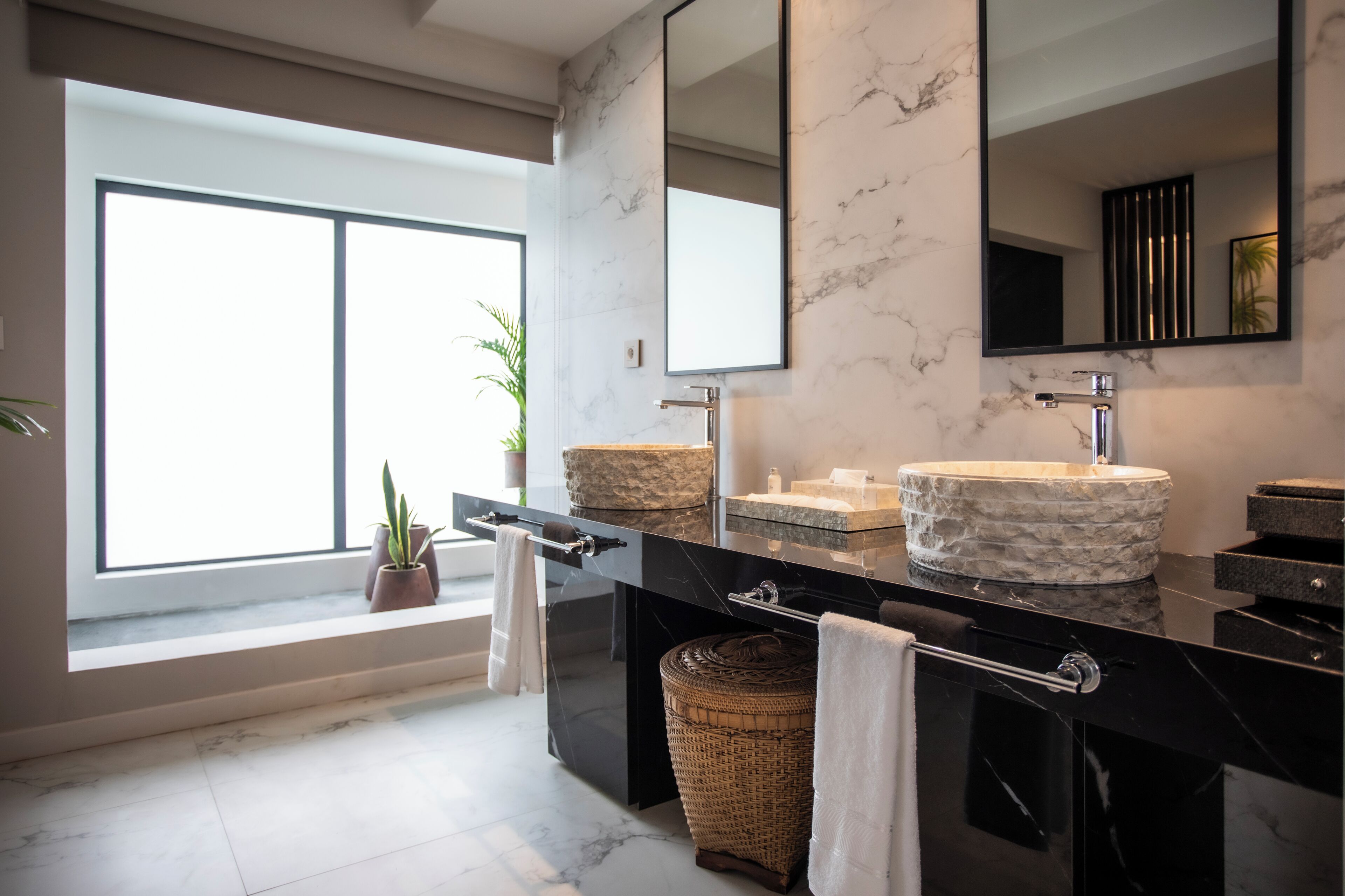 Un baño moderno con un ventanal grande, dos lavamanos con encimeras de mármol, un espejo grande y una cesta de mimbre.
