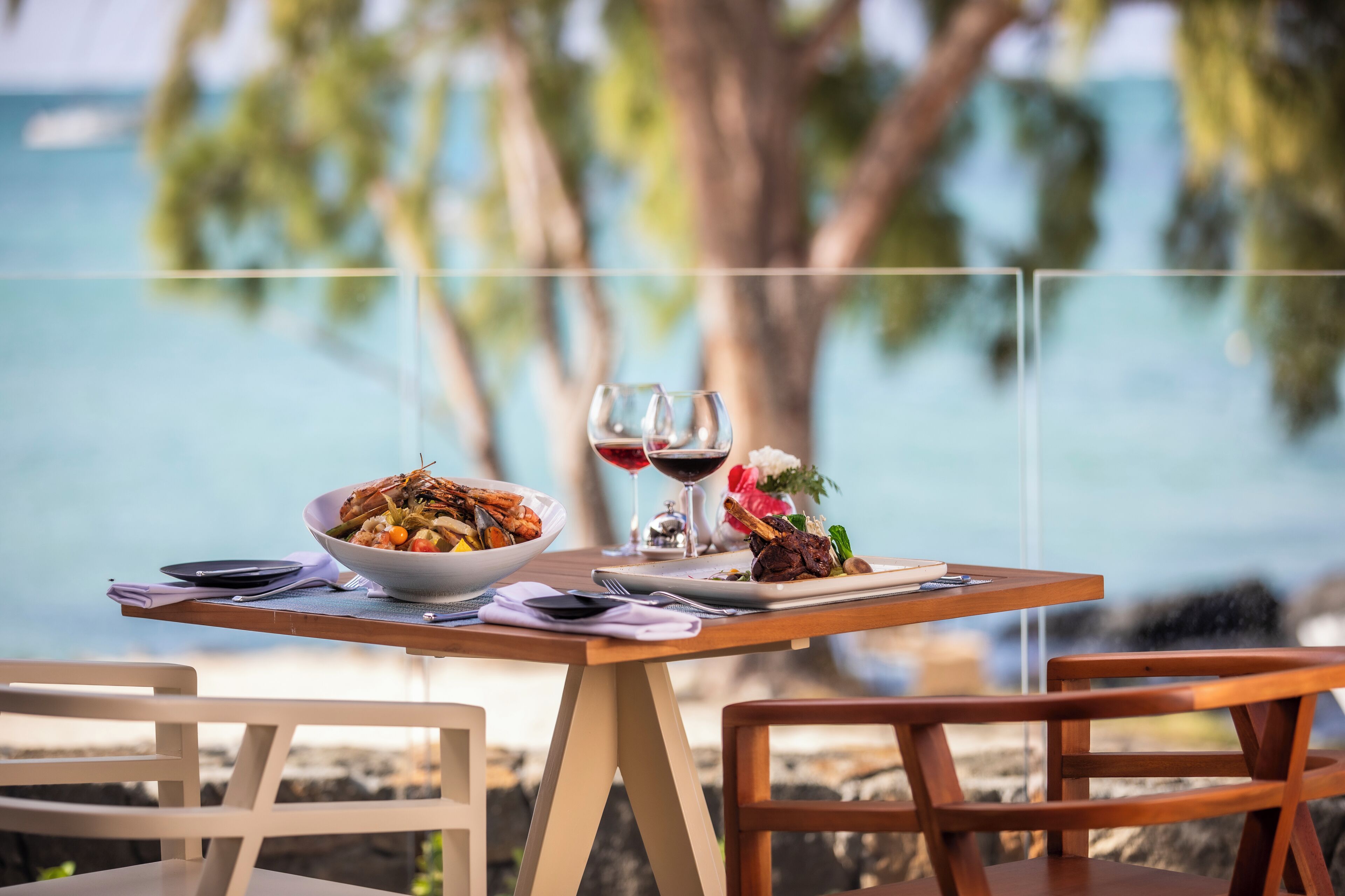 A dining table with a bowl of food, two wine glasses, and a plate of food, set against a scenic view of a body of water and trees.