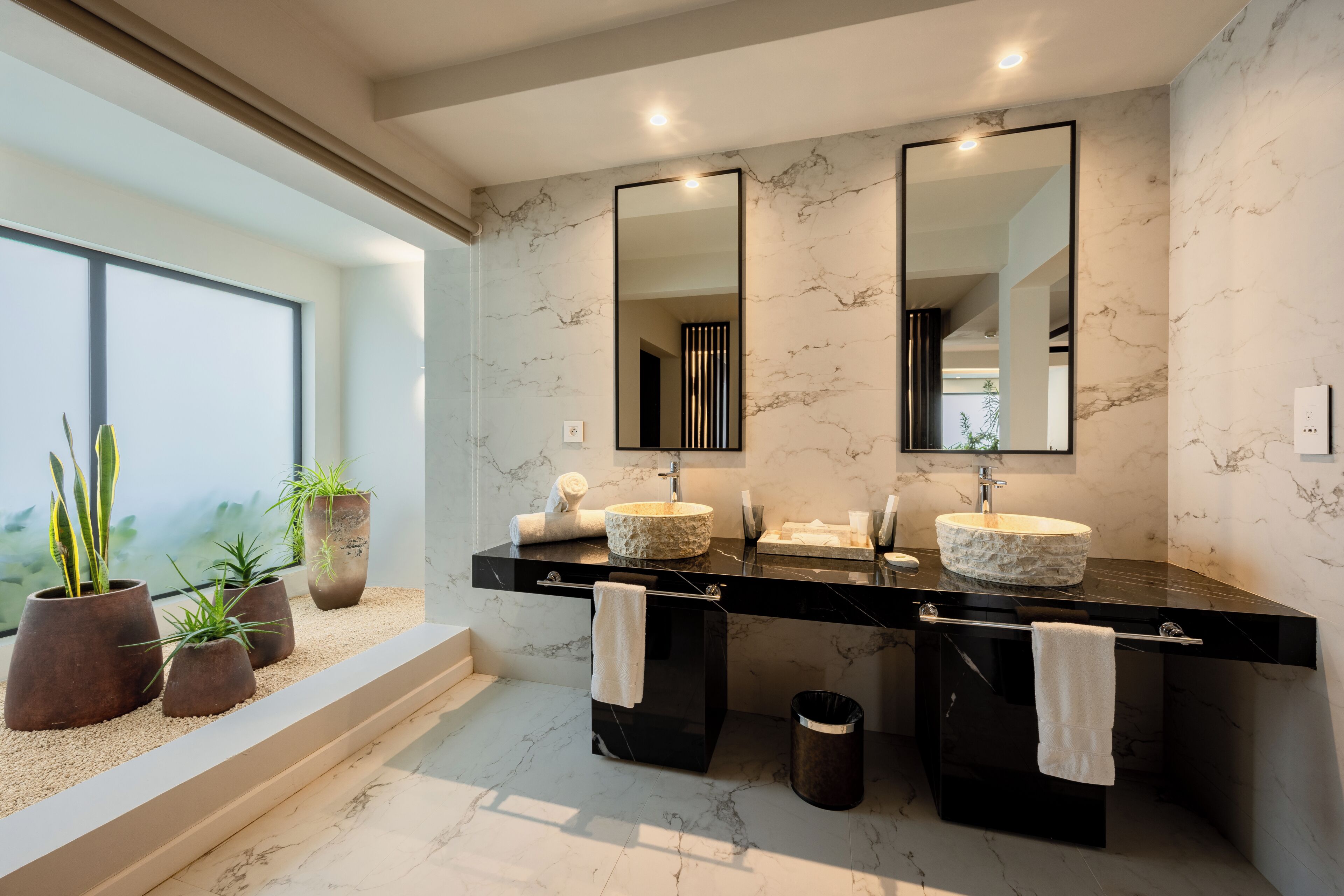 Un baño moderno con dos lavamanos, paredes de mármol y plantas en macetas.