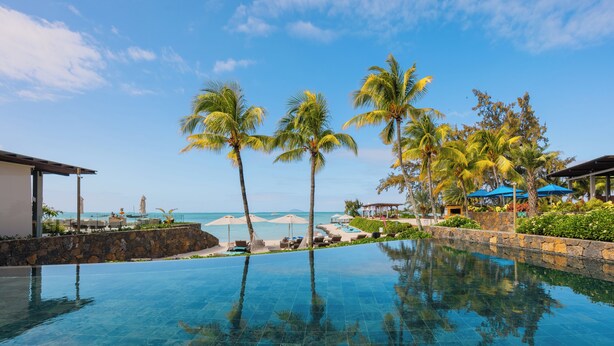 A pool with clear blue water, surrounded by palm trees and a stone wall, with a view of the ocean and beach umbrellas. - Sea Diamond Boutique Hotel & Spa by Sea Resorts (Calodyne)