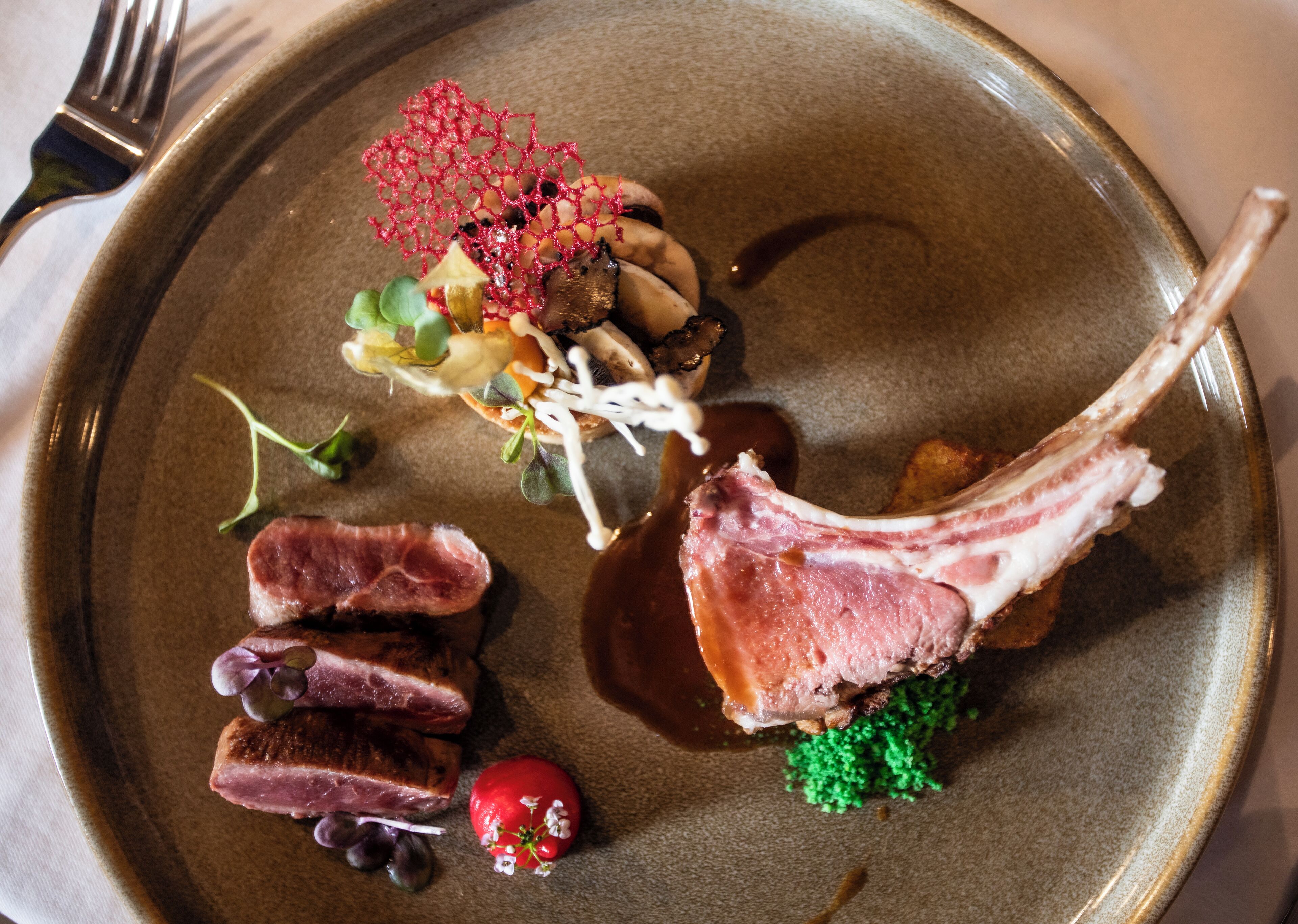 Un plato con diversos alimentos, incluyendo un chuletón de cordero, setas y una estructura roja decorativa.
