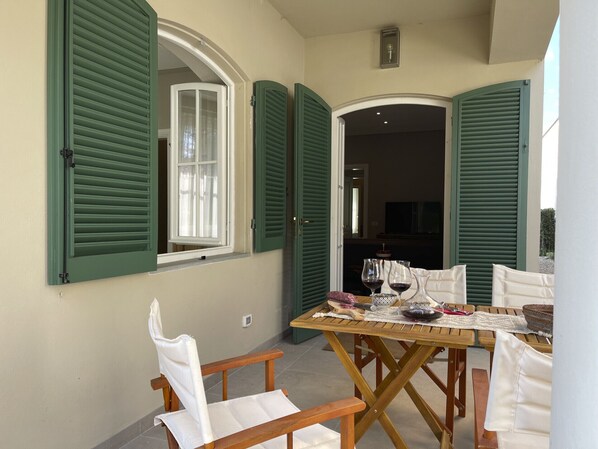 A dining area with a wooden table and chairs, a window with green shutters, and a view into the adjacent room.