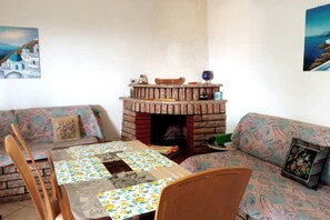 A cozy living room with a brick fireplace, patterned sofas, a dining table with a floral tablecloth, and two chairs.