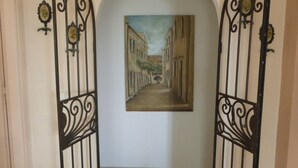 Un couloir orné d’un tableau représentant une scène de rue, doté d’un sol carrelé et flanqué de deux grilles noires à motifs.